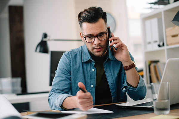 young man in casual attire talking on the phone while working at a desk with a laptop and papers focused on task productivity and organization in a modern workspace